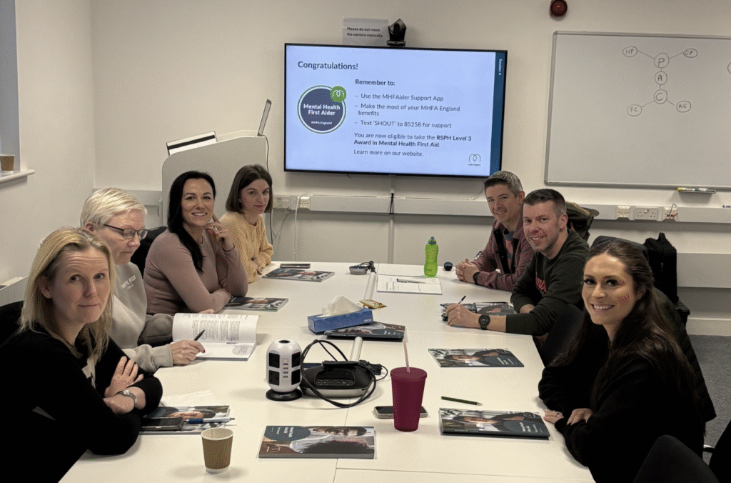 A group of people smile at the camera all seated at a large white desk in a small conference room