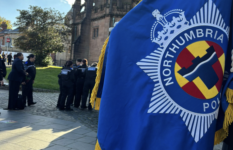 A blue flag with the Northumbria Police logo on it flies with police officers in the background