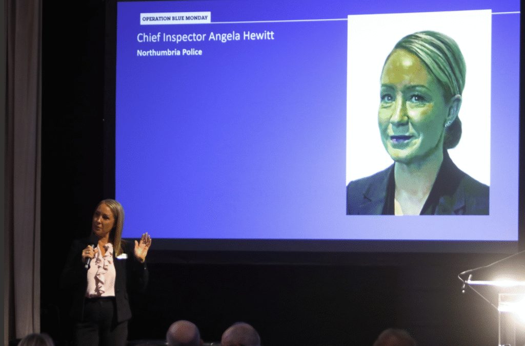 A lady wearing a white top and black suit addresses an audience on stage with a picture of her face behind her.