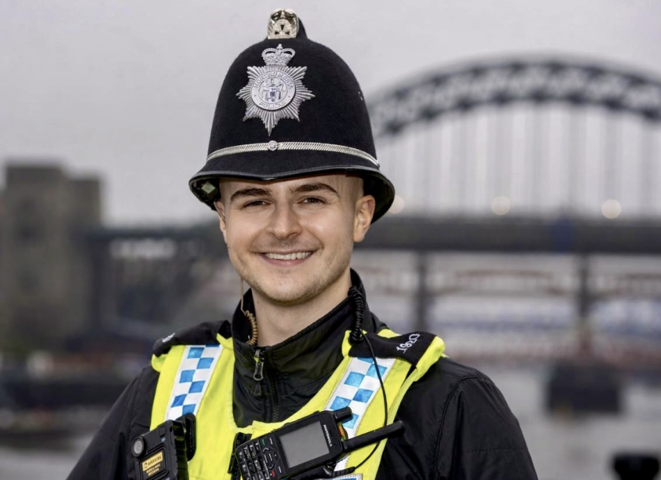 PC Tom Kelly wears full police uniform and smiles in front of a big bridge in the background